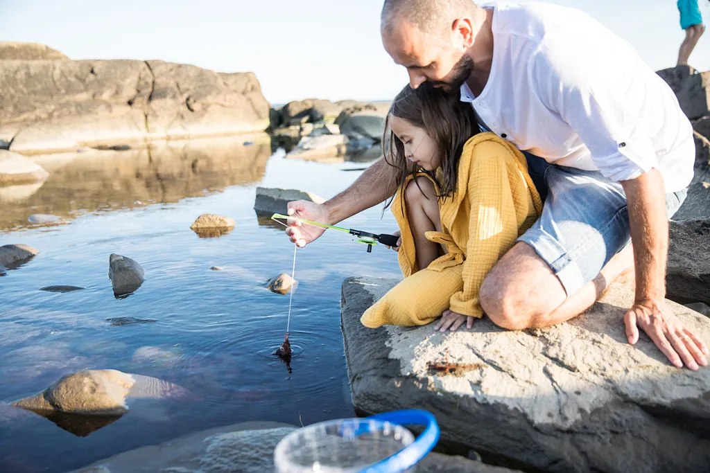 En pappa och dotter som sitter på stenar vid havet och fiskar krabbor.