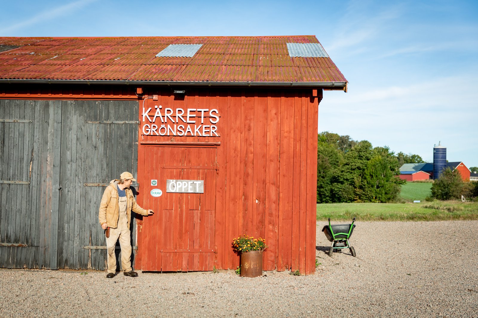 En röd ladugård där en man står framför och håller i dörrhandtaget, ovanför står det "Kärrets grönsaker".