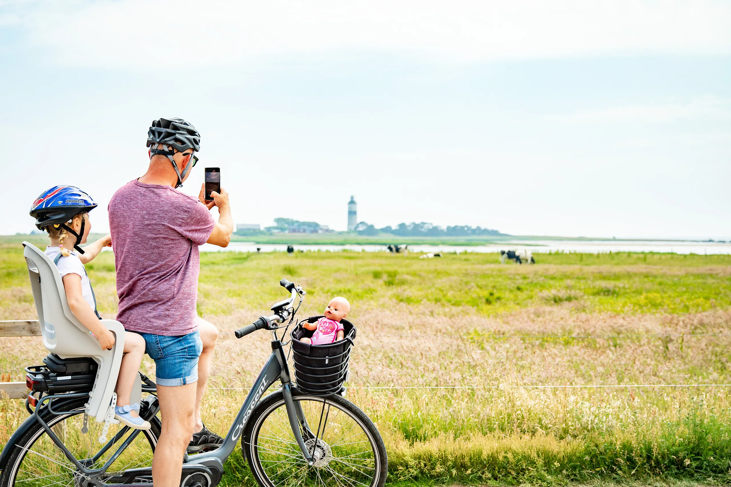 En man har stannat sin cykel och tar ett kort med mobilen på en fyr och ett strandlandskap. Dottern sitter cykelsadel.
