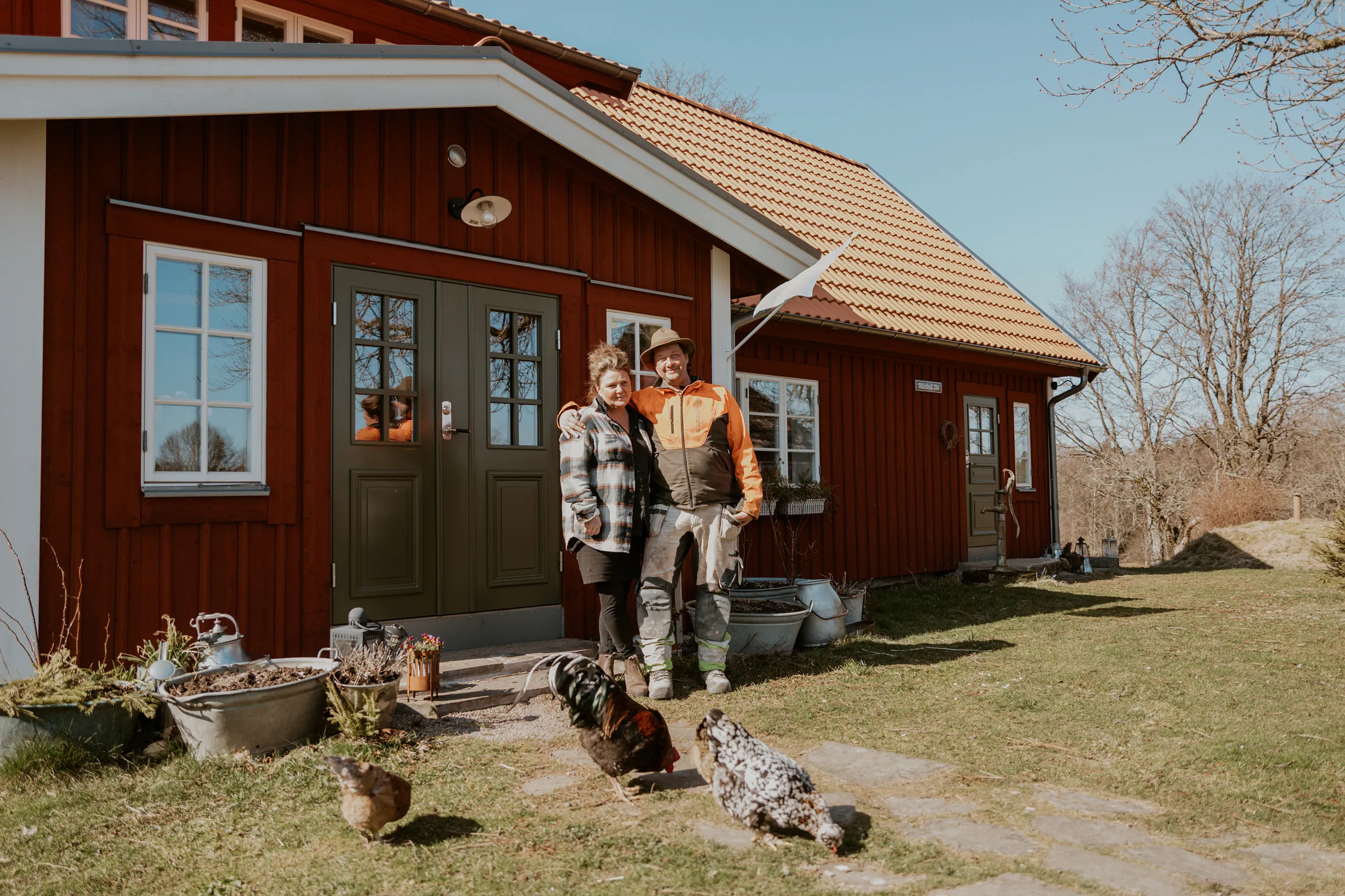 Två personer står framför ett rött hus med hönor vid sina fötter