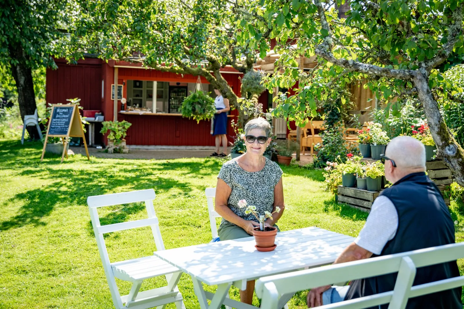 Kvinna som sitter i ett vitt möblemang i ett trädgårdscafé.