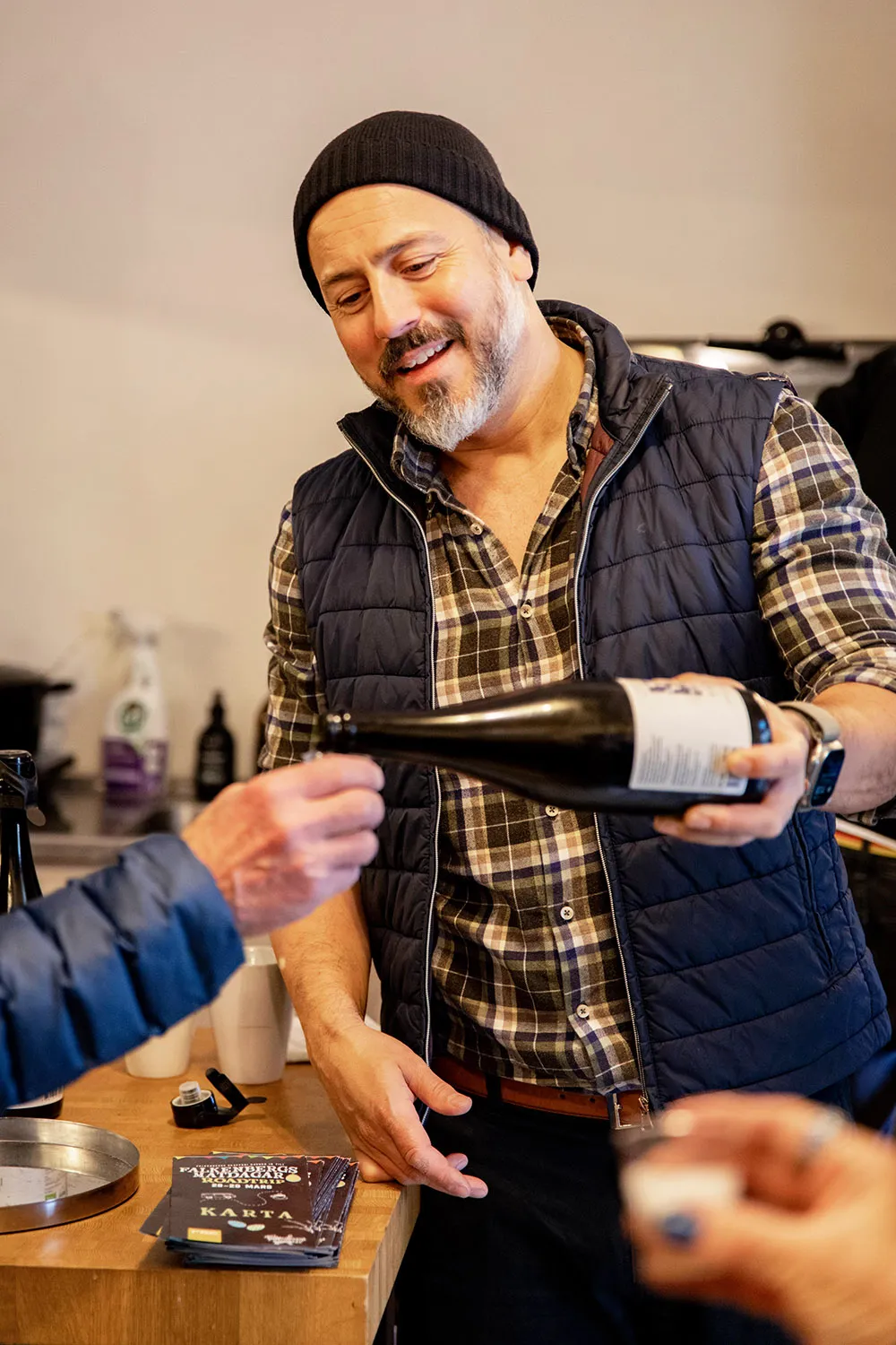 En man med skjorta.väst och mössa, häller upp ur en flaska i ett provglas som någon håller fram.